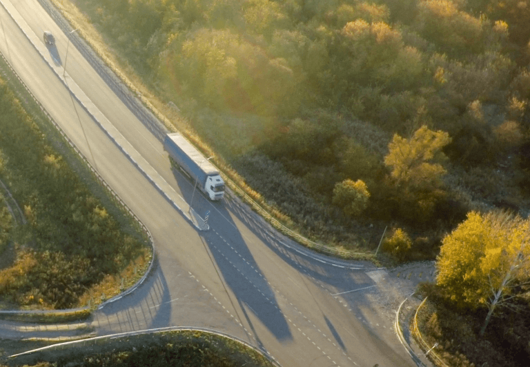 Truck approaching four-way intersection of four-lane highway with trees on either side of the highway.