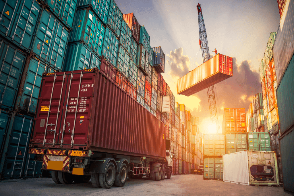 A crane moving shipping container in a shipyard with containers staked high on both sides and a truck with a shipping container waiting.