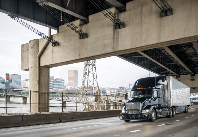 Truck traveling on bridge under another bridge with a city in the background.