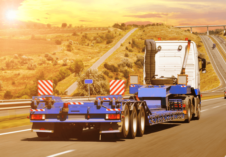 An empty flatbed truck on a highway with sand and sparse plants on either side.