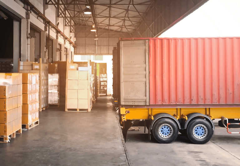 loading dock laden with pallets of boxes with a trailer backed up to receive.