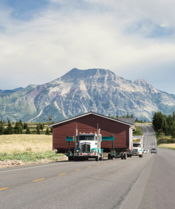 Green and white tractor hauling a heavy brown cabin down a road with mountains in the background.
