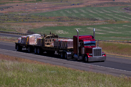 Red tractor with flat bed hauling two trailers ful of different freight.