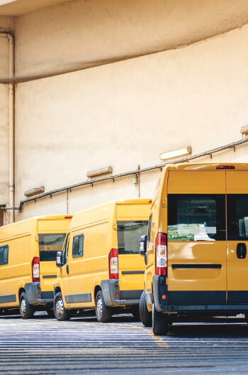 Several yellow sprinter vans line up behind a warehouse.