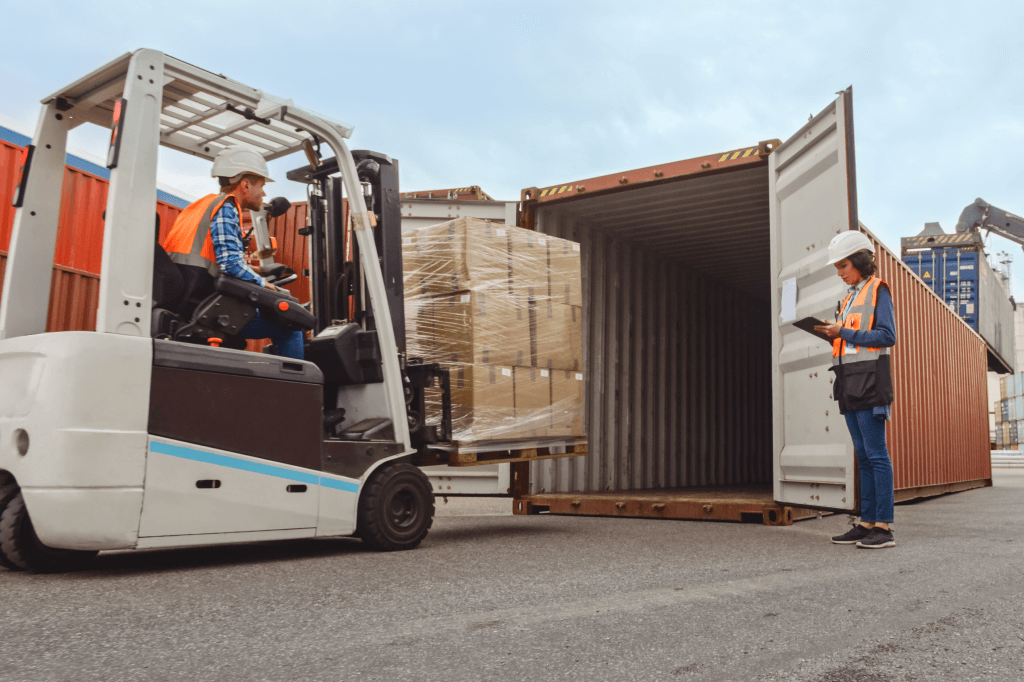 A person driving a forklift, loading a pallet onto an open shipping container with a person supervising.